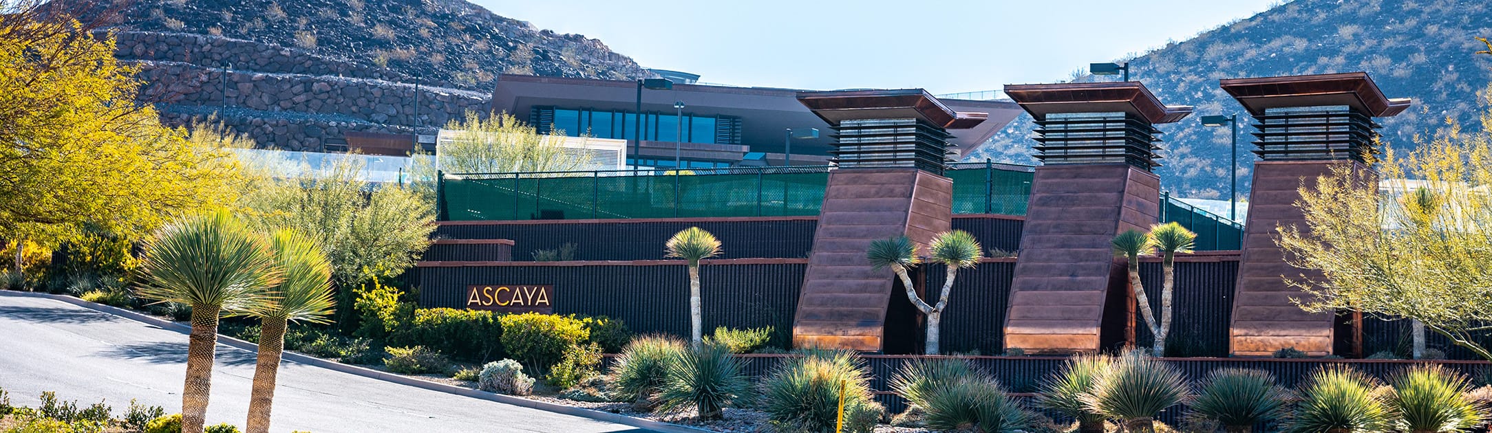 3 black layered stands with sign on bottom layer that says Ascaya. 3 brick pillars with lighting on top and street entrance to community.