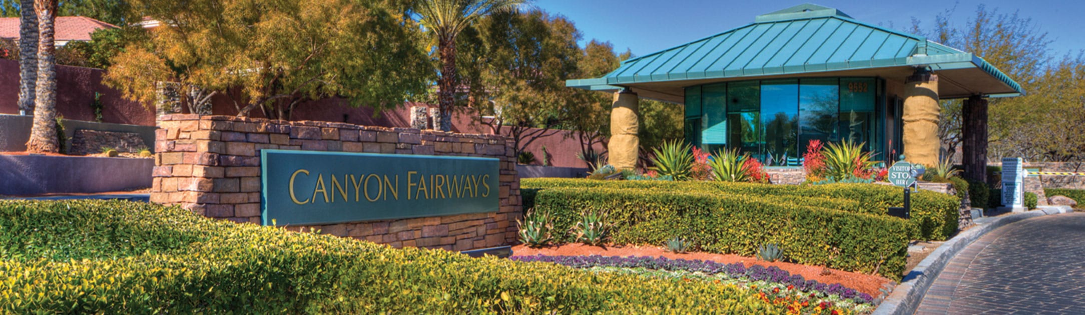 Brick stand with green sign that says Canyon Fairways, bushes, birds of paradise plants and gate entrance building behind.