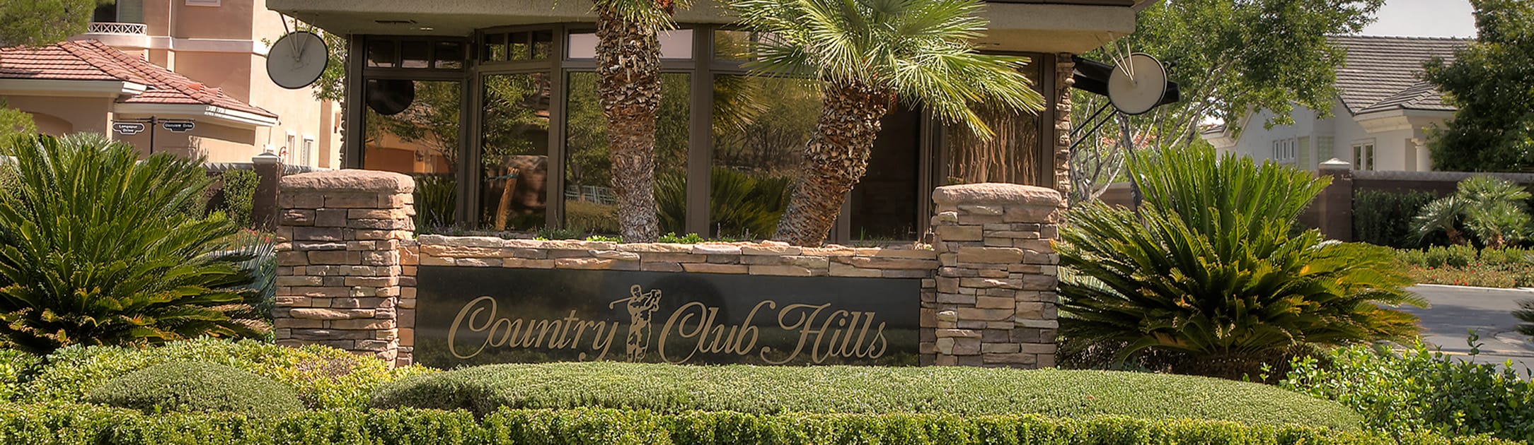 Brick stand with sign that says Country Club Hills, bushes, palm trees and gate entrance building behind.