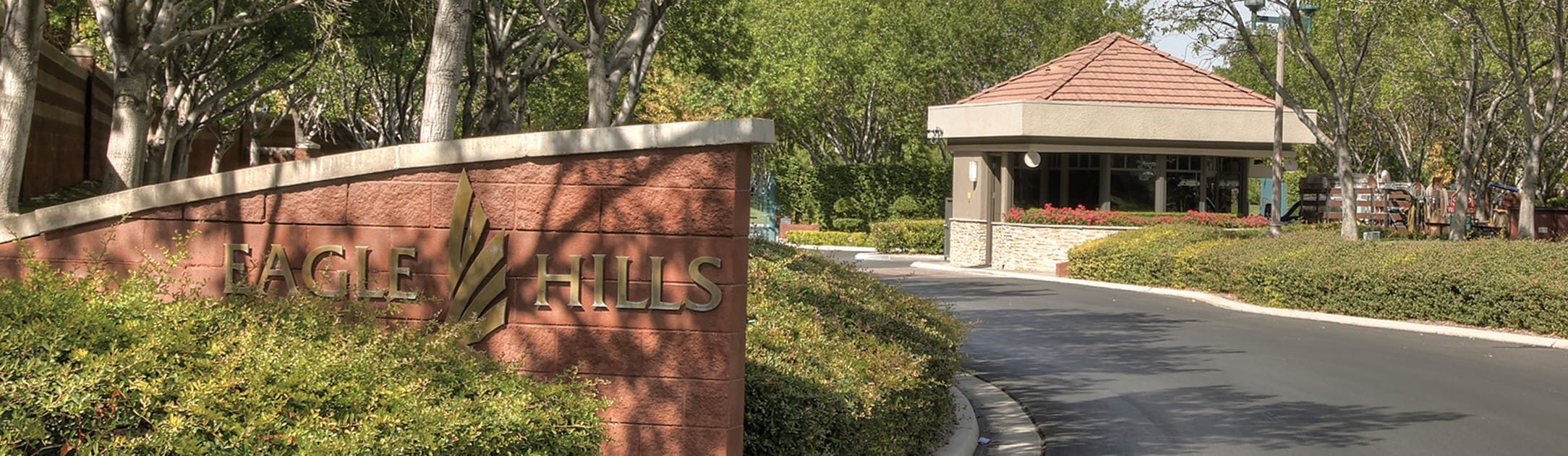 Brick stand with sign that says Eagle Hills, bushes, trees and gate entrance building behind.