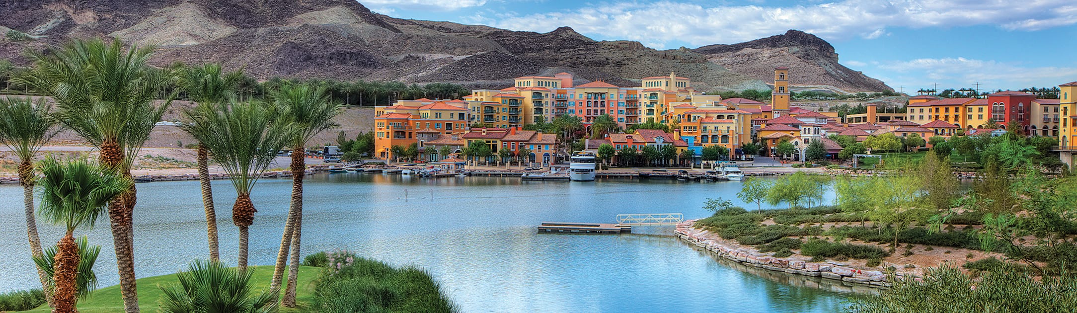 Golf course view of lake with color building that are different heights and mountains in the background.