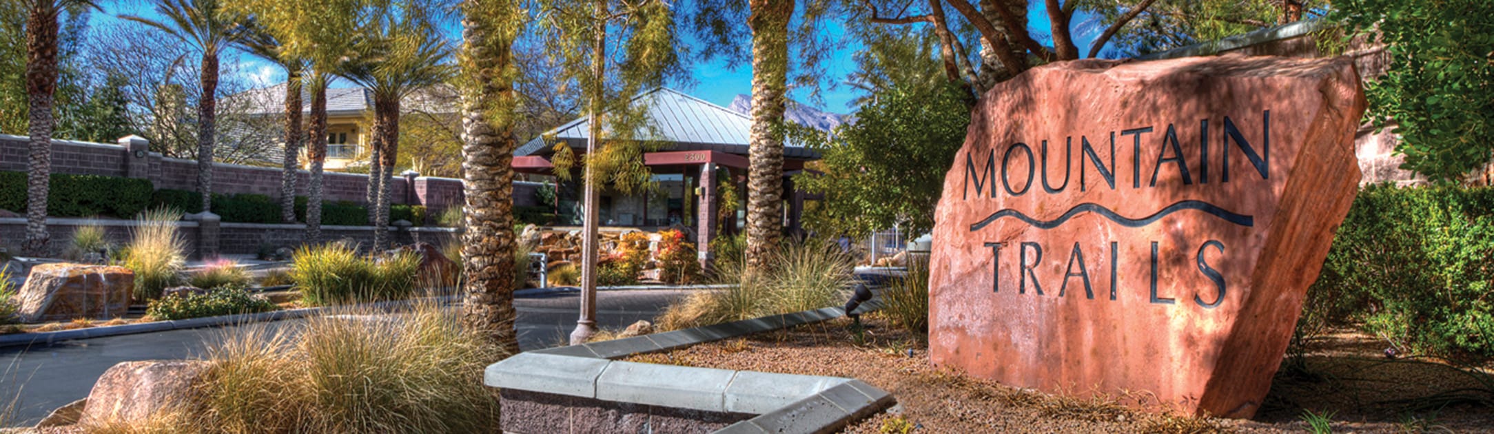 Entrance to Mountain Trails community with red rock stone and name engraved in it with trees and road.