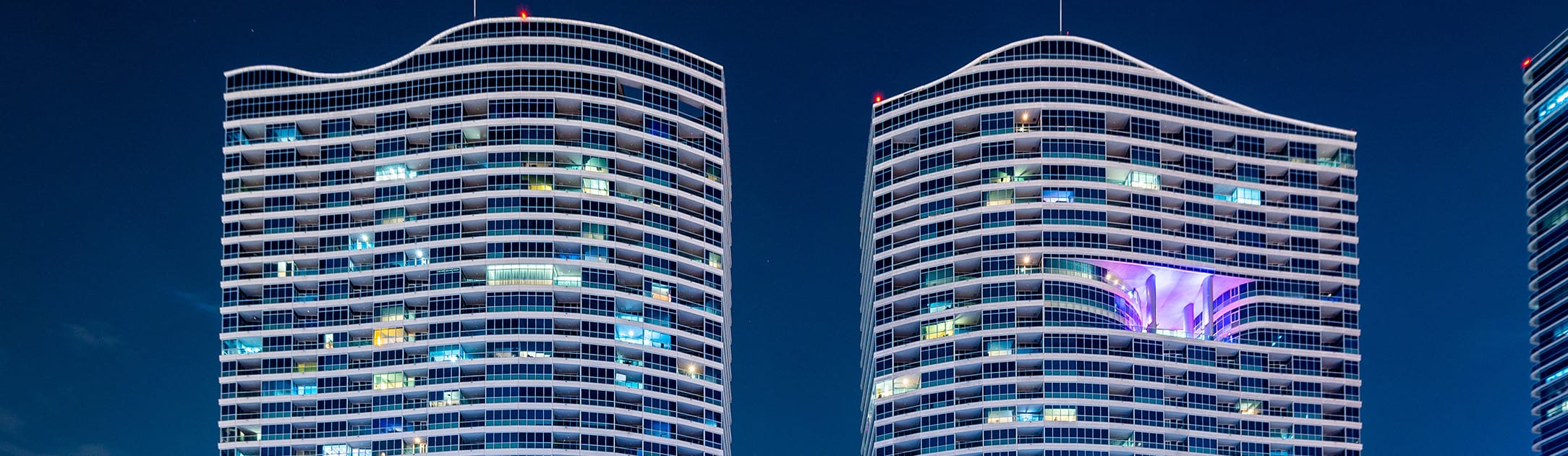 Top view of 2 high rise towers at night with white, blue and purple lights.