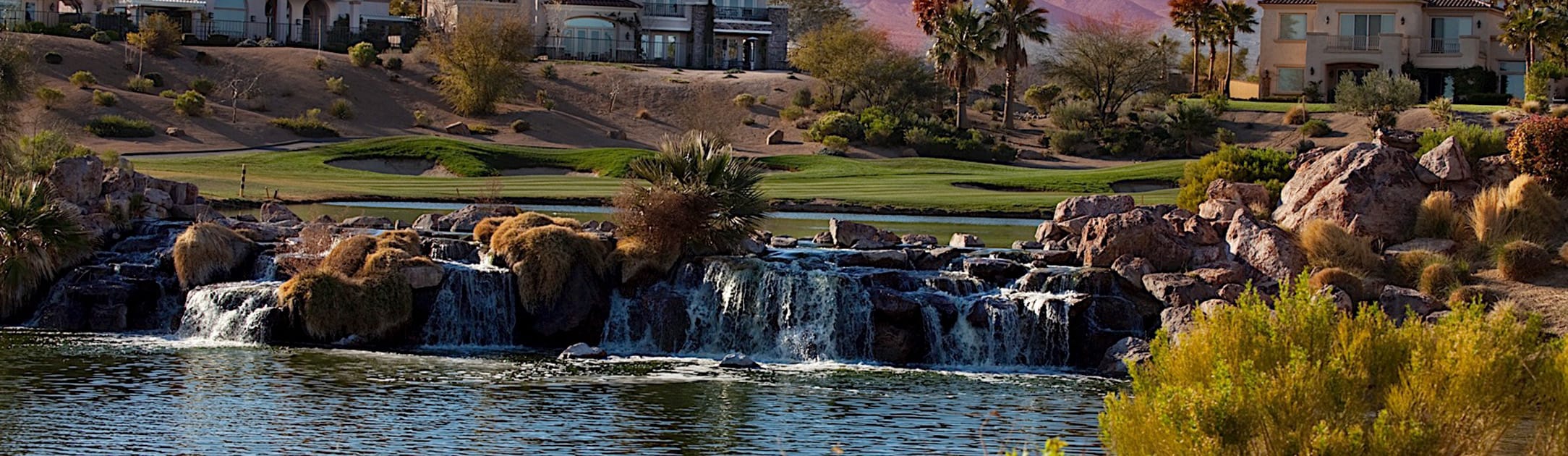 Rock waterfall with golf course, lake, palm trees and homes behind it.