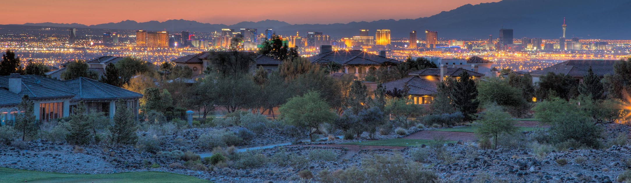 Twilight aerial view of golf course and neighborhood with high rises and mountains in the background with orange sunset.