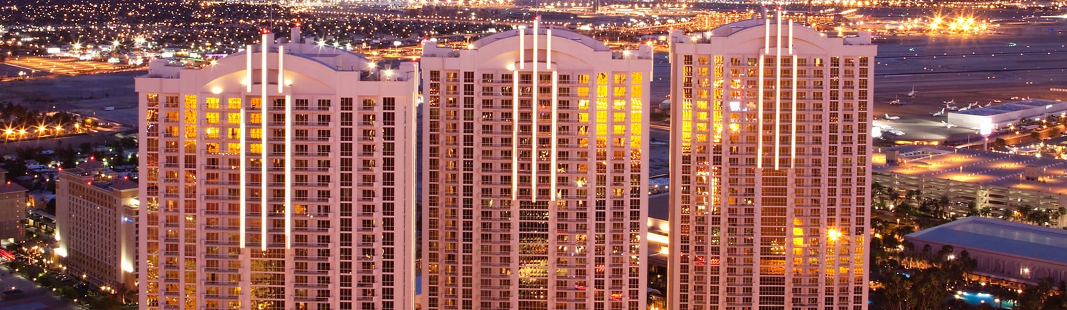 Night view of the top of 3 high rises with pink and yellow hues. High rises and businesses in the background.
