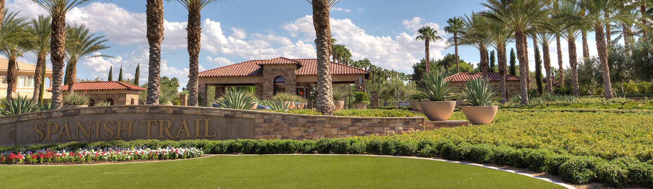 Brick stand with sign that says Spanish Trails, grass, flowers, bushes, palm trees and gate entrance building behind.