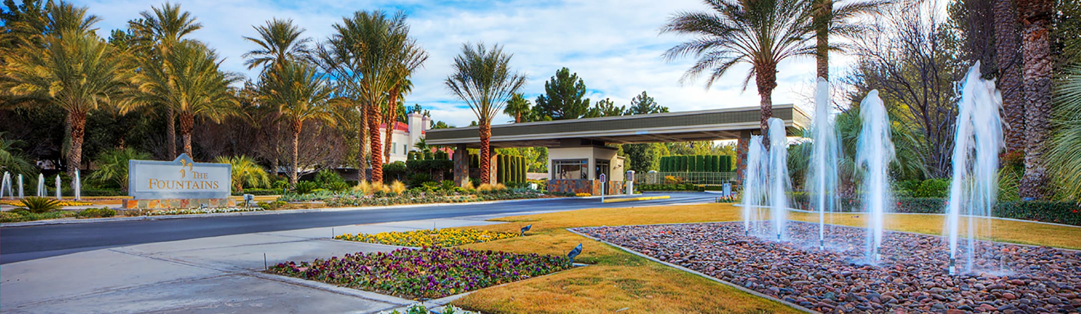 Flowers, grass and 5 fountains in red rock bed in front. Stone sign that says the Fountains with road to gate entrance building with palm trees in the back.