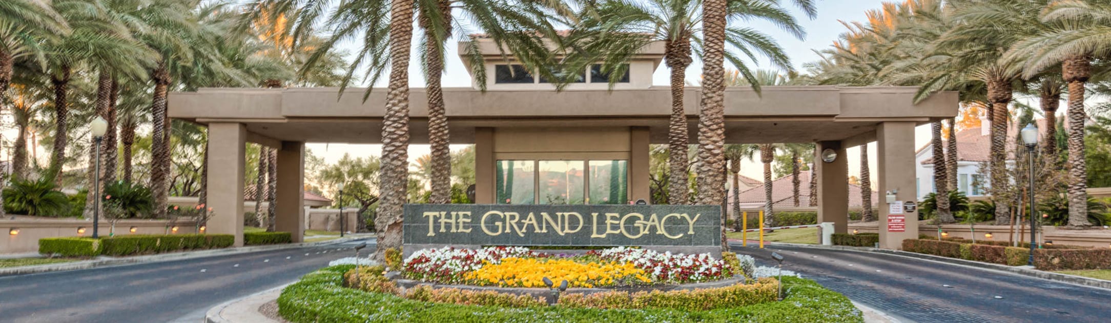 Brick stand with sign that says The Grand Legacy, flowers, bushes, palm trees and gate entrance building behind.