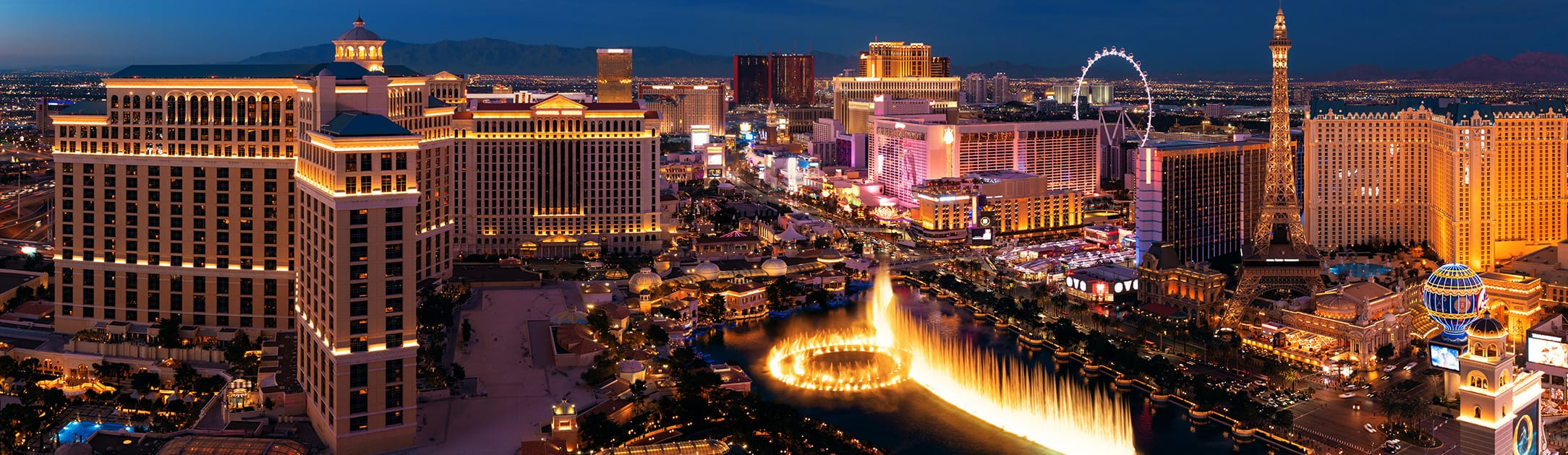 Night time view of hotels, casinos, water fountain, ferris wheel and in the background are homes and mountains.