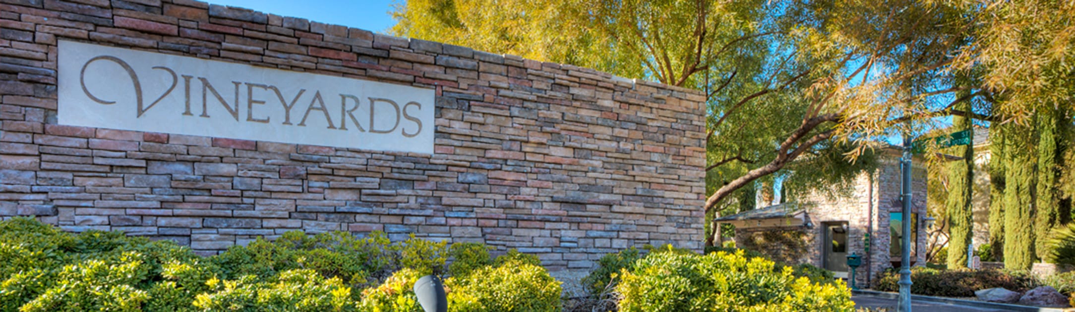 Brick stand with sign that says Vineyards, bushes, trees and gate entrance building behind.