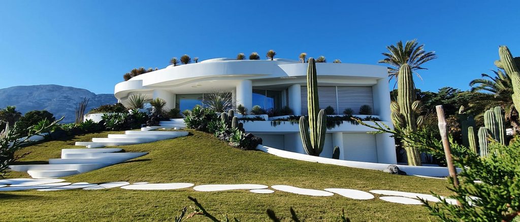 White oval house with a flat roof, circle stone steps that go up a hill ad around the house and palm trees.