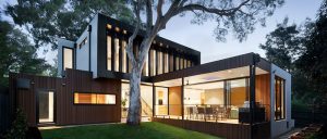 Night time shot of a 2 story modern home with walls of glass with lights on and trees outside.