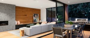 Living room with gray brick fireplace, wood panel wall, couch, wall of glass views and dining room behind it.