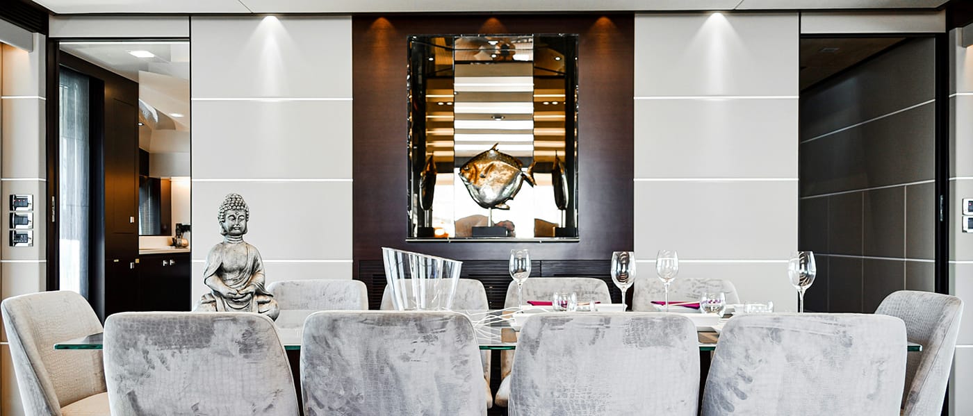 Dining room with lush gray chairs, Budda, wine glasses and decanter on table. Glass fish ornament and glass shelves in nook on the wall.