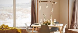 Dining table sat with wine glasses, tall candles on candle sticks, tree branch across the ceiling with lights hanging from it and window with view of snow.